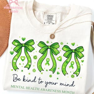 May include: Cream-colored t-shirt with three green bows and the text "Be kind to your mind" and "Mental Health Awareness Month." The bows have heart and ribbon details. A gold necklace with a pendant is draped on the shirt.
