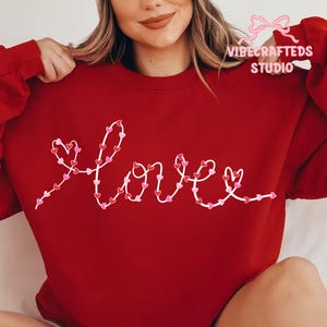 Puede incluir: Una sudadera roja con la palabra "Love" bordada en blanco, con corazones rosas y rojos. La sudadera tiene mangas largas y cuello redondo. El texto "VIBECRAFTEDS STUDIO" está en la esquina superior derecha.