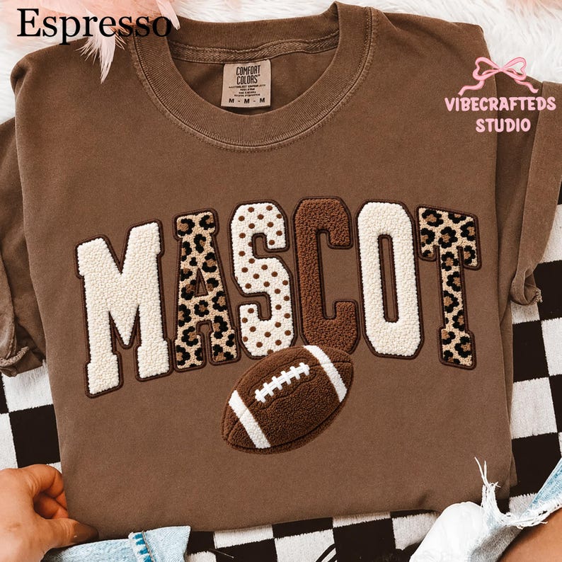 May include: A brown t-shirt featuring the word MASCOT in textured, multi-patterned varsity-style letters. The letters alternate between cream, leopard print, and brown. Below the text is a brown and white textured football graphic.