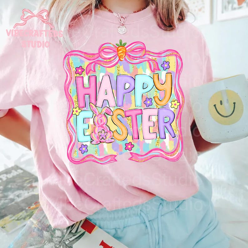 May include: A light pink t-shirt with a colorful "Happy Easter" graphic. The graphic features a bunny, flowers, and a carrot on a pink frame. The shirt is worn by a person holding a white mug with a yellow smiley face.