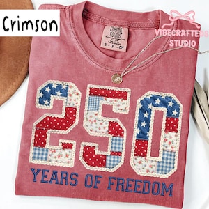 May include: Crimson-colored t-shirt with the number "250" in a patchwork design featuring red, white, and blue patterns. The text "YEARS OF FREEDOM" is below the number. A silver necklace with a pendant is on the shirt.