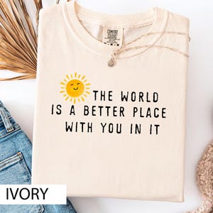 May include: An ivory crew neck t-shirt featuring a yellow sun graphic with a smiling face and the black text, THE WORLD IS A BETTER PLACE WITH YOU IN IT. The shirt has a Comfort Colors brand label at the inner neckline and a short sleeve design.