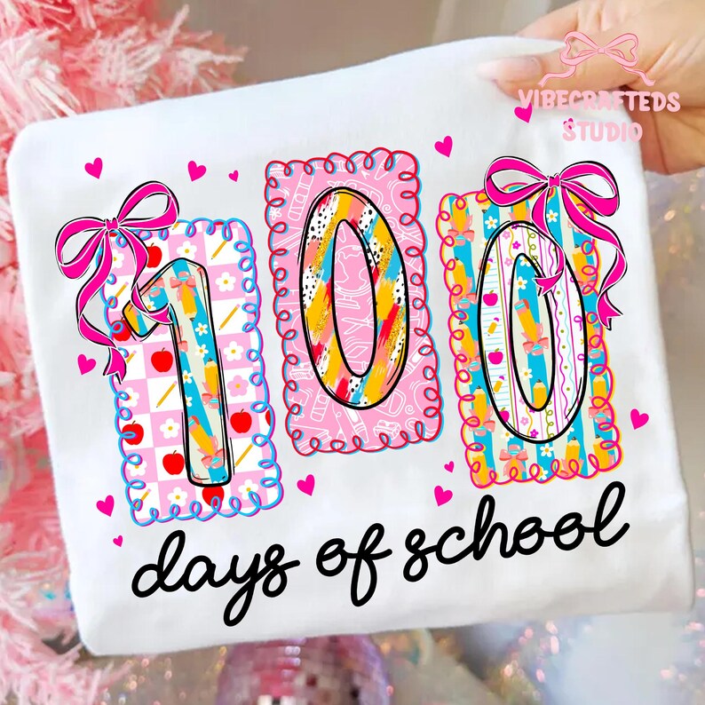 May include: White t-shirt with a colorful "100 days of school" graphic. The design features the numbers "100" in a playful font, decorated with bows, hearts, and school-themed patterns. The text "days of school" is written in a cursive font.