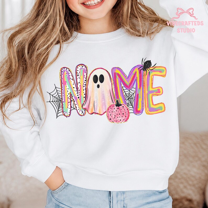 May include: A white long-sleeve sweatshirt featuring a colorful graphic design with the word NAME, where the letter A is replaced by a ghost, accompanied by a pink polka-dot pumpkin, black spiderwebs, and a small black spider on the letter M.