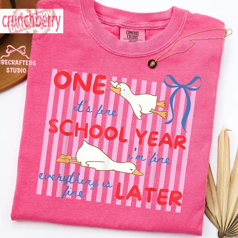 May include: A pink t-shirt with the text "ONE school year later" and the phrase "everything is fine". The shirt features a graphic of two white cartoon geese. A gold necklace with a pendant is visible.