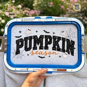 Puede incluir: Una tela gris claro sujeta en un bastidor de bordado azul de 20x33 cm con el texto negro brillante PUMPKIN y el texto naranja en cursiva season. El diseño incluye figuras de fantasmas blancos, murciélagos negros, telarañas y estrellas.