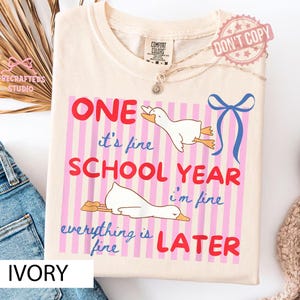 May include: Ivory-colored t-shirt with a graphic featuring a duck and the text "ONE it's fine SCHOOL YEAR i'm fine everything is fine LATER." The design includes pink stripes and a blue bow.