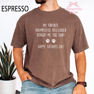 May include: Espresso-colored t-shirt with white text that reads "MY FAVORITE UNEMPLOYED FREELOADER BOUGHT ME THIS SHIRT" with paw prints and "HAPPY FATHER'S DAY". The shirt is a short-sleeved, crew neck style.