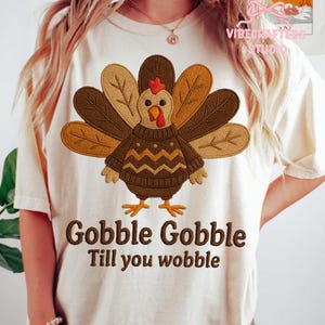 Könnte beinhalten: Cremefarbenes T-Shirt mit einem detaillierten, gestickten Truthahndesign. Der Truthahn trägt einen Pullover und hat braune, beige und orangefarbene Federn. Der Text "Gobble Gobble Till you wobble" steht darunter.