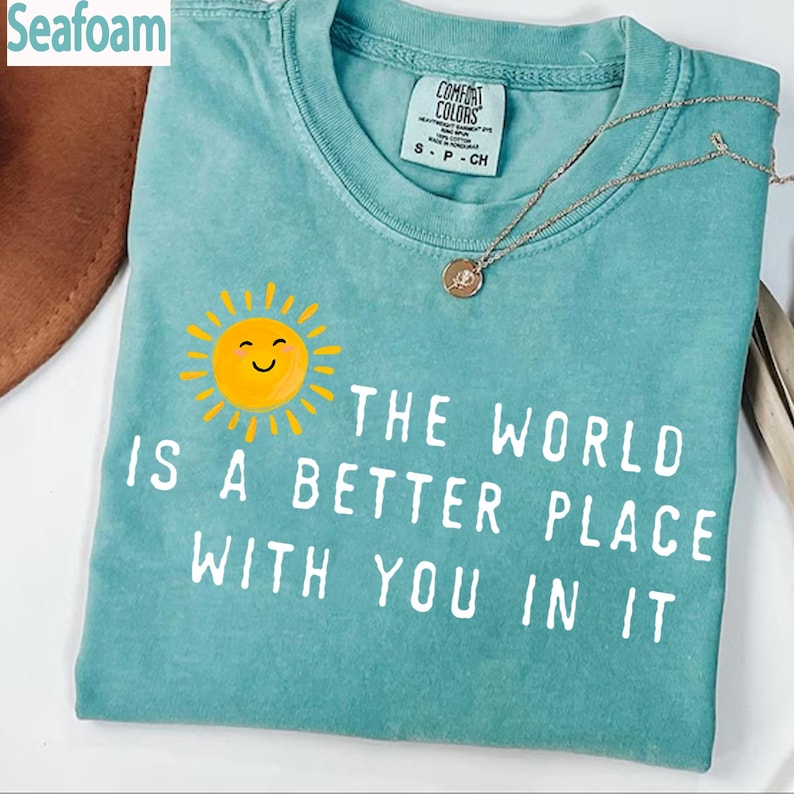 May include: A seafoam green short-sleeve t-shirt featuring a yellow smiling sun graphic and the white text "THE WORLD IS A BETTER PLACE WITH YOU IN IT". The shirt has a crew neckline and a Comfort Colors brand label on the inside collar.