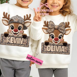 May include: Two white sweatshirts featuring reindeer designs. One reindeer wears a cap and holds a sign with "KEVIN". The other has a bow and holds a sign with "OLIVIA". A pink sticker reads "custom name".