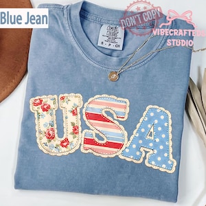 Puede incluir: Camiseta azul claro con la palabra "USA" aplicada en la parte delantera. Las letras están decoradas con patrones florales, de rayas y de lunares. Un collar dorado está drapeado sobre la camiseta.