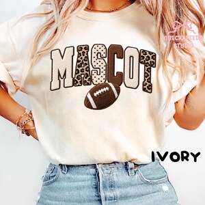 May include: An ivory t-shirt featuring the word MASCOT in large, textured block letters with varying patterns including leopard print, polka dots, and solid brown. A brown football graphic is positioned below the text. The word IVORY is printed.