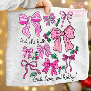 May include: A light gray sweatshirt featuring a collection of pink bows with various patterns and designs. The text "Deck the halls with bows and holly!" is printed in a script font, creating a festive holiday theme.
