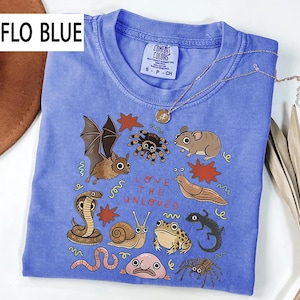 May include: Flo Blue Comfort Colors t-shirt featuring a graphic of a bat, spider, mouse, and snake. The text "LOVE THE UNLOVED" is printed on the front. A silver necklace with a pendant is draped over the shirt.