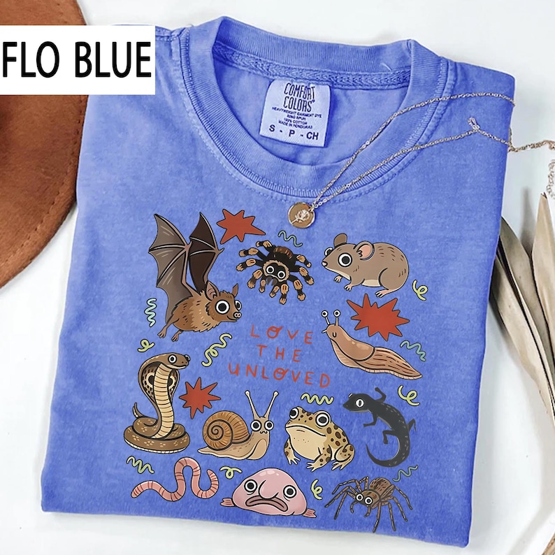 May include: Flo Blue Comfort Colors t-shirt featuring a graphic of a bat, spider, mouse, and snake. The text "LOVE THE UNLOVED" is printed on the front. A silver necklace with a pendant is draped over the shirt.