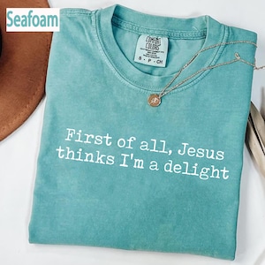 Puede incluir: Camiseta verde mar con el texto blanco "First of all, Jesus thinks I'm a delight". La camiseta tiene cuello redondo y una etiqueta Comfort Colors. Un collar dorado con un colgante descansa sobre la camiseta.