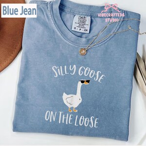 May include: A blue jean colored t-shirt with the words "Silly Goose On The Loose" and a graphic of a goose wearing sunglasses. The shirt has a crew neck and short sleeves. The Comfort Colors brand tag is visible.