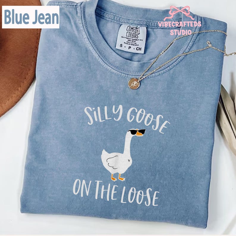 May include: A blue jean colored t-shirt with the words "Silly Goose On The Loose" and a graphic of a goose wearing sunglasses. The shirt has a crew neck and short sleeves. The Comfort Colors brand tag is visible.
