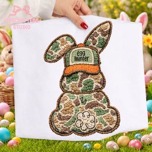 May include: White fabric with an embroidered Easter bunny design. The bunny is in a camouflage pattern with a green and orange cap that reads "egg hunter". The design is held by a person with a red manicure.
