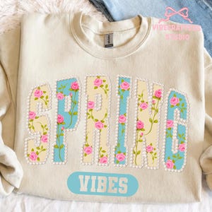 May include: Cream-colored sweatshirt with the word "SPRING" in a floral design. The letters are outlined with pearls and filled with a light blue and cream pattern, adorned with pink roses. The word "VIBES" is printed below.