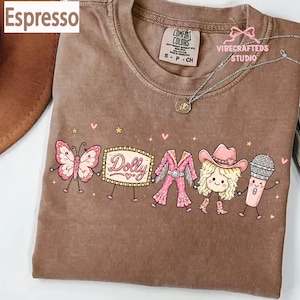 May include: An espresso-colored t-shirt featuring a graphic print of a butterfly, a marquee sign with the text Dolly, a pink fringed outfit, a blonde character wearing a cowboy hat, and a microphone, all holding hands with small stars and hearts.
