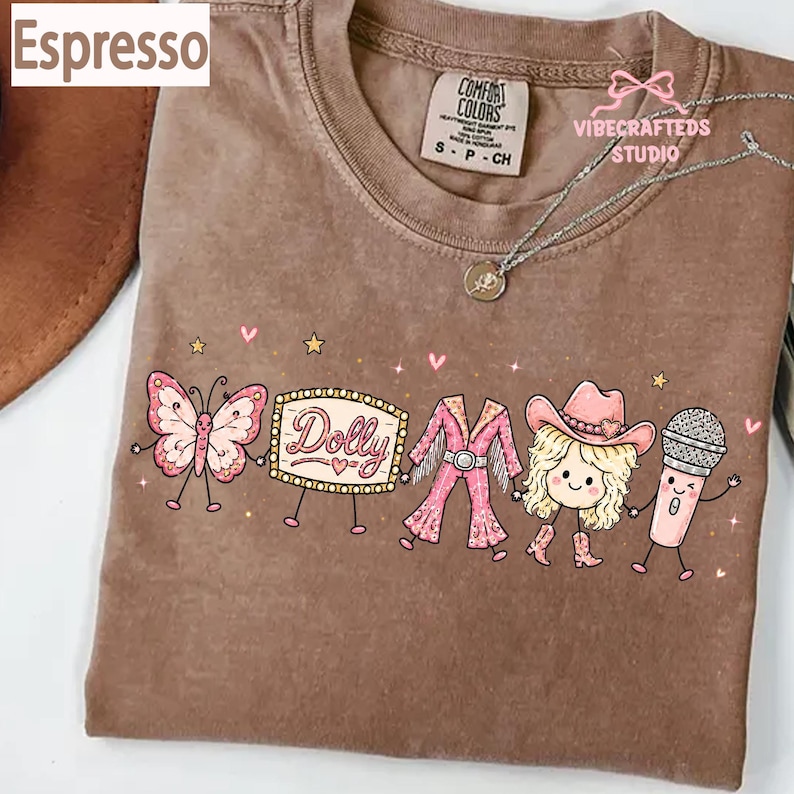 May include: An espresso-colored t-shirt featuring a graphic print of a butterfly, a marquee sign with the text Dolly, a pink fringed outfit, a blonde character wearing a cowboy hat, and a microphone, all holding hands with small stars and hearts.