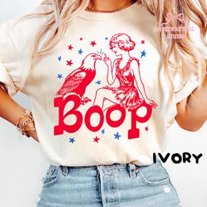 May include: Ivory t-shirt with a red graphic of an eagle and a woman, and the word "Boop" in red. Blue and red stars surround the design. The word "IVORY" is printed in black.