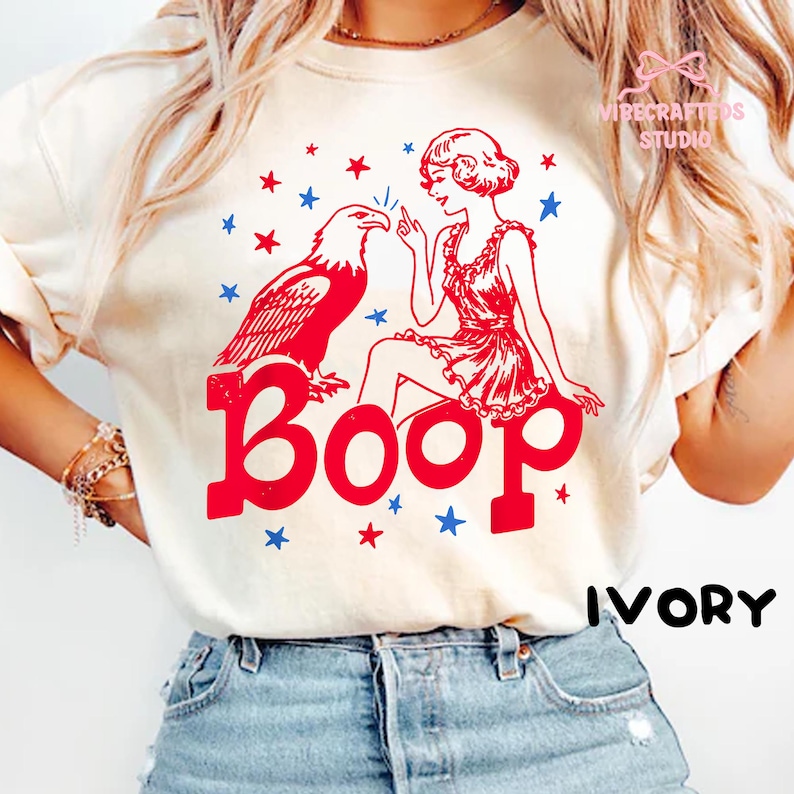 May include: Ivory t-shirt with a red graphic of an eagle and a woman, and the word "Boop" in red. Blue and red stars surround the design. The word "IVORY" is printed in black.