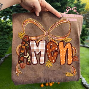 Puede incluir: Un artículo de tela marrón con la palabra "AMEN" en un diseño con temática otoñal. Las letras están decoradas con estampados florales, cuadros y lunares, con un gran lazo en la parte superior y hojas de otoño.