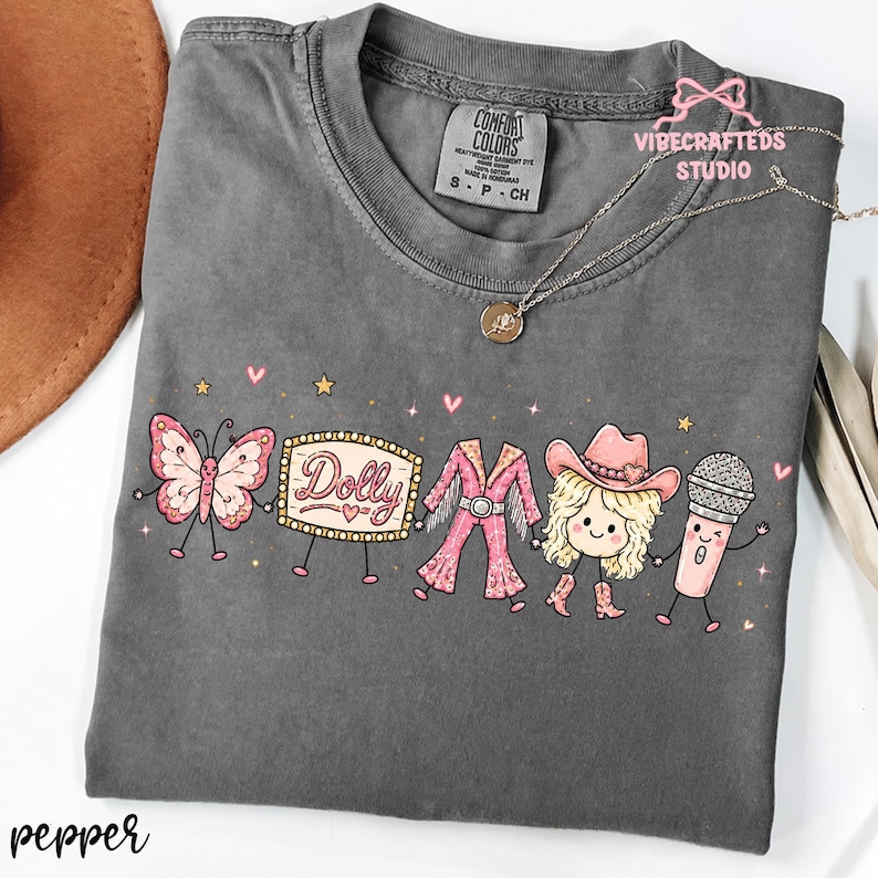May include: A charcoal gray short-sleeve t-shirt featuring a graphic print of a pink butterfly, a marquee sign with the text Dolly, a pink fringe outfit, a blonde character in a cowboy hat, and a microphone, all with arms and legs.