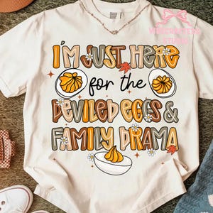 Può includere: T-shirt color crema con la scritta "I'M JUST HERE for the DEVILED EGGS & FAMILY DRAMA" in lettere marroni e arancioni. Il design include illustrazioni di uova farcite, margherite e foglie autunnali.