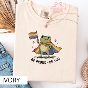 Pode incluir: Camiseta de cor marfim com um gráfico de um sapo usando uma capa de arco-íris e segurando uma bandeira de arco-íris. O texto "BE PROUD BE YOU" está abaixo do sapo. A camisa tem uma etiqueta Comfort Colors.