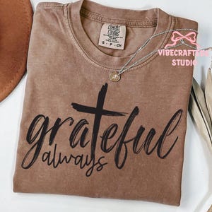 May include: A brown Comfort Colors t-shirt with the words "grateful always" in black script and a black cross. A silver necklace with a small pendant rests on the shirt. The shirt is folded neatly.