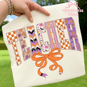 May include: Cream-colored sweatshirt with the word "TEACHER" in orange, purple, and black block letters. The letters have various patterns, including checkerboard, chevron, and Halloween-themed graphics. An orange bow is at the bottom.