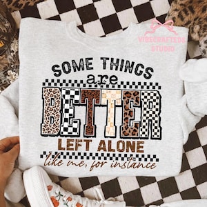 Puede incluir: Sudadera gris claro con la frase "SOME THINGS are BETTER LEFT ALONE like me, for instance." La palabra "BETTER" está diseñada con estampado de leopardo y patrones de cuadros. Sudadera de estilo casual.
