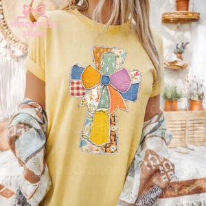 May include: Yellow t-shirt featuring a patchwork cross design with a large bow. The cross is made of various fabric patterns in shades of blue, orange, pink, and yellow. The bow is multi-colored. The shirt has short sleeves.
