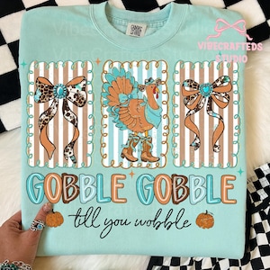 Può includere: Una felpa azzurra con tema del Ringraziamento. Presenta un tacchino con cappello e stivali da cowboy, affiancato da fiocchi decorativi. Il testo "GOBBLE GOBBLE till you wobble" è visualizzato sotto.