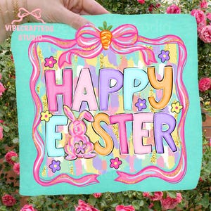 May include: A turquoise square with the words "HAPPY EASTER" in colorful bubble letters. A pink bunny and flowers decorate the design, which is framed by a pink ribbon and bow with a carrot accent. The words "VIBECRAFTED STUDIO" are in the upper left corner.