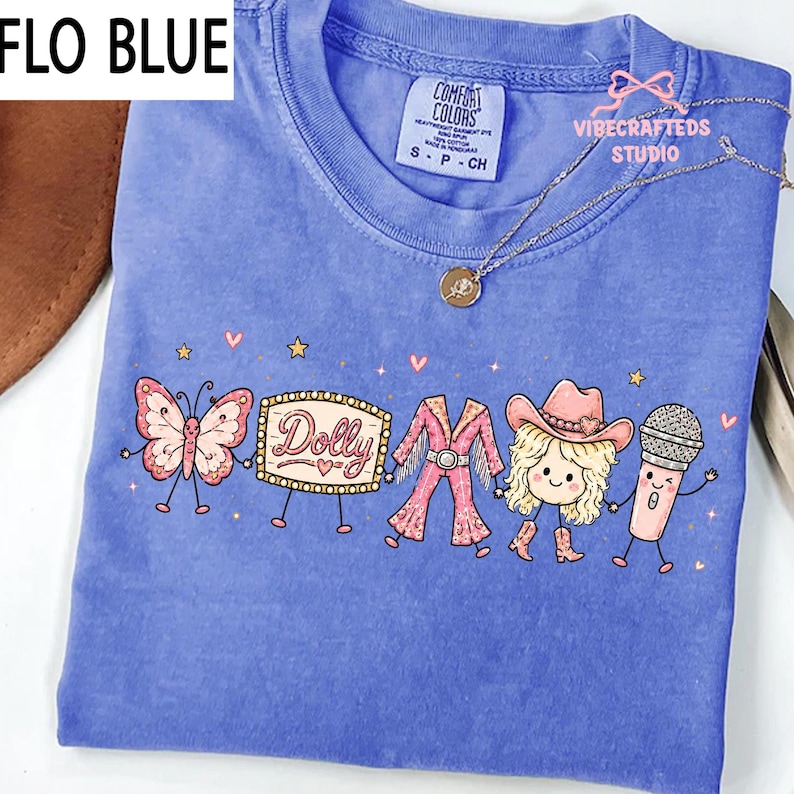May include: A periwinkle blue t-shirt featuring a graphic print of a butterfly, a marquee sign with the text Dolly, a pink fringe outfit, a blonde character wearing a cowboy hat and boots, and a microphone, all holding hands with small stars.