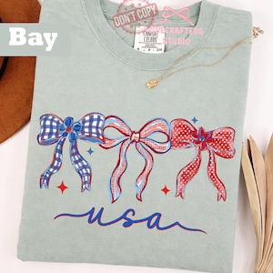 May include: A pale green Comfort Colors t-shirt with three patriotic bows in red, white, and blue. The bows are hand-painted with gingham and polka dot patterns. The word "usa" is written in blue script below the bows.