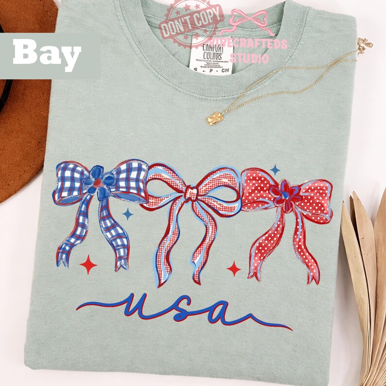 May include: A pale green Comfort Colors t-shirt with three patriotic bows in red, white, and blue. The bows are hand-painted with gingham and polka dot patterns. The word "usa" is written in blue script below the bows.