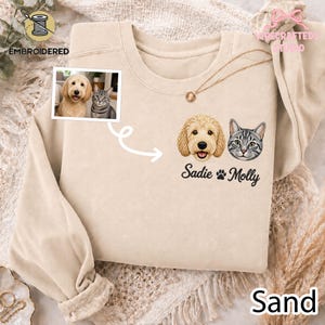 Può includere: Una felpa girocollo color sabbia con un ricamo personalizzato raffigurante un golden doodle e un gatto tigrato. Sotto i ritratti degli animali, i nomi Sadie e Molly sono ricamati in corsivo nero, separati da un'icona a forma di zampa.
