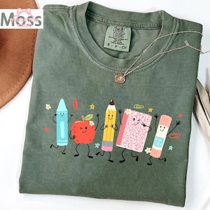 Puede incluir: Una camiseta de manga corta color verde oliva con un estampado gr&aacute;fico de cinco &uacute;tiles escolares antropom&oacute;rficos: un cray&oacute;n azul, una manzana roja, un l&aacute;piz amarillo, un cuaderno rosa y una barra de pegamento, todos con caras sonrientes.