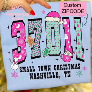 May include: Light blue t-shirt with the text "SMALL TOWN CHRISTMAS NASHVILLE, TN" and a custom ZIPCODE design. The design includes pink and green numbers, snowflakes, a snowman, gingerbread man, Christmas tree, and a mug.