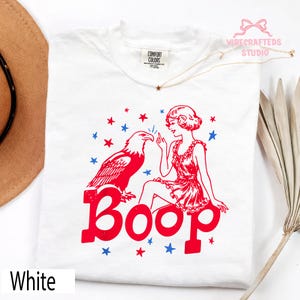 May include: White t-shirt with a red and blue graphic. The design includes a red eagle, a woman in a red dress, and the word "Boop" in red. Blue and red stars are scattered around the design. The text "White" is at the bottom.