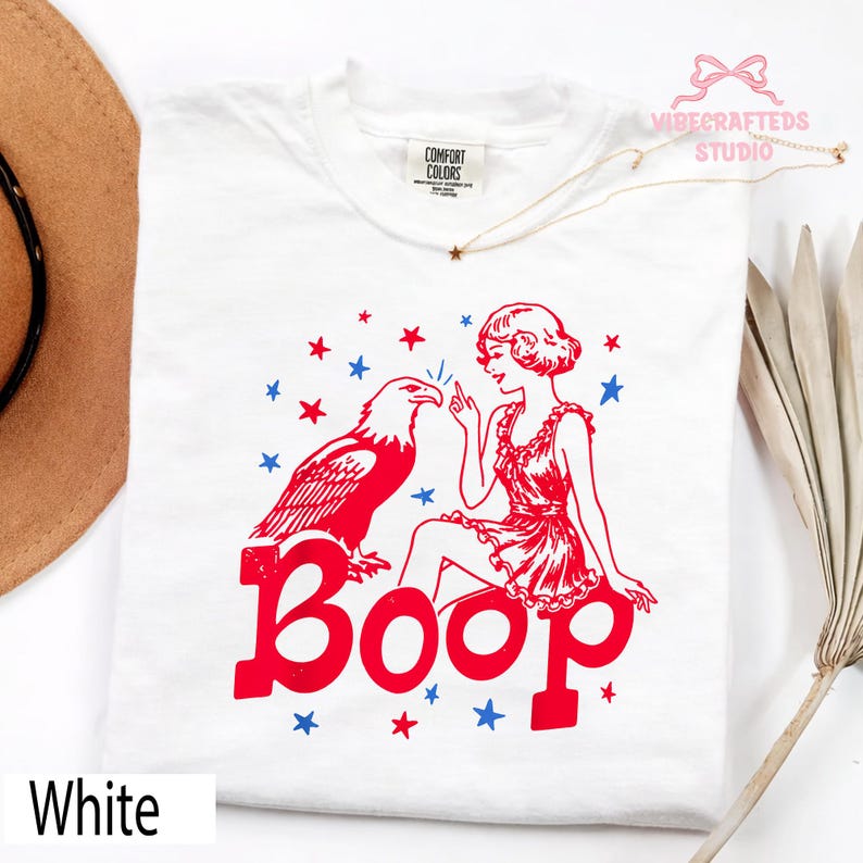 May include: White t-shirt with a red and blue graphic. The design includes a red eagle, a woman in a red dress, and the word "Boop" in red. Blue and red stars are scattered around the design. The text "White" is at the bottom.