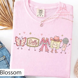 May include: A light pink t-shirt featuring a graphic print of a butterfly, a marquee sign with the text Dolly, a pink fringe outfit, a blonde character wearing a cowboy hat and boots, and a microphone, all holding hands with small hearts and stars.