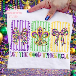 Op de afbeelding: Wit T-shirt met een kleurrijk Mardi Gras-ontwerp. Het shirt heeft drie rechthoekige kaders met een strik, fleur-de-lis en strik ontwerp. De tekst "LET THE GOOD TIMES ROLL" staat eronder.