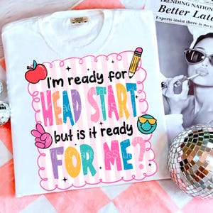 May include: White t-shirt with a colorful graphic that reads "I'm ready for HEAD START but is it ready FOR ME?" The design includes an apple, pencil, peace sign, and a smiling face with sunglasses. The shirt is on a pink and white checkered surface.